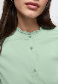 Blusa verde menta con colletto arricciato, cinque bottoni sul davanti e un tessuto morbido e texturizzato.