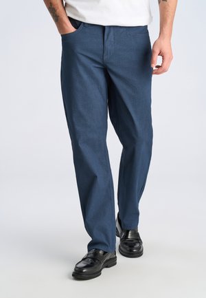 Mężczyzna w niebieskich teksturowanych spodniach, czarnych skórzanych mokasynach i białej koszuli, z ręką w kieszeni, stojący na jasnoszarym podłożu.