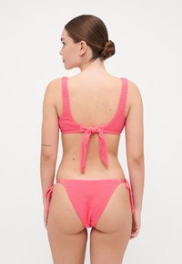 Roza teksturirana bikinijska postava z zgornjim delom s širokimi naramnicami in zavezovanjem na hrbtu ter nastavljivimi spodnjimi hlačkami s vezanjem ob strani, ki poudarjajo gladko obrabo.