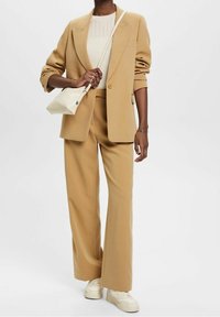 Beżowa, dopasowana marynarka z szerokimi nogawkami, beżowe spodnie do kompletu oraz lekki kremowy sweter. Do tego mała biała torebka na ramię.