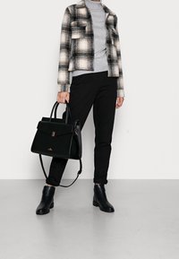 Svart läderhandväska med strukturerad design, guldfärgad detaljer och dubbla handtag. Modellen är klädd med en rutig jacka, grå polotröja och svarta byxor.