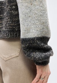 Χέρι ακουμπισμένο στο πλάι, φορώντας πουλόβερ με χοντρή πλέξη σε σκούρα και ανοιχτά χρώματα και μπεζ παντελόνι με  ορατές ραφές στην πίσω τσέπη.