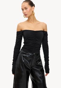 Vrouw in een zwarte off-shoulder top met ruches en lange mouwen, gedragen met hoge zwarte nep-leer broek met krokodillenpatroon, en accessoires van grote gouden creolen.