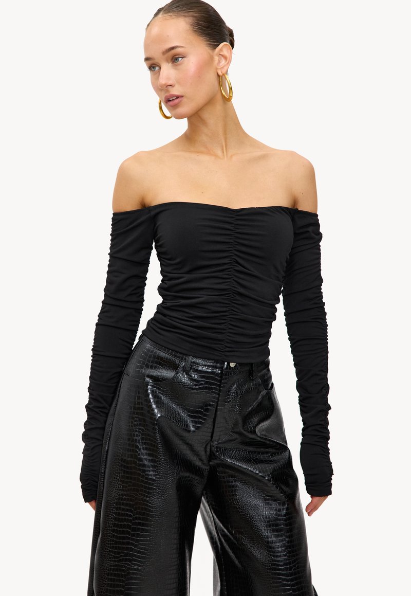 Vrouw in een zwarte off-shoulder top met ruches en lange mouwen, gedragen met hoge zwarte nep-leer broek met krokodillenpatroon, en accessoires van grote gouden creolen.