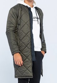 Quiltet olivengrøn dynejakke med lang pasform, med sort lynlås foran og sidelommer, lagdelt over en hvid hættetrøje.