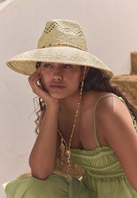Pălărie din paie cu boruri largi, cu un model țesut și accente din mărgele multicolore, asortată cu o rochie verde cu pliuri. Materiale naturale, texturate.