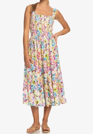 Femme portant une robe midi sans manches à motif floral avec un corsage froncé et des sandales, debout avec une main sur la hanche.