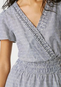 Žena odjevena u plavo-bijelu prugastu haljinu na preklop s naboranim rubom i elastičnim pojasom, prikazujući gornji torzo i ruku.