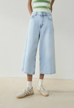 Personne portant un jean large bleu clair coupé, un haut sans manches rayé, des chaussettes blanches et des baskets beiges, debout sur un sol uni.