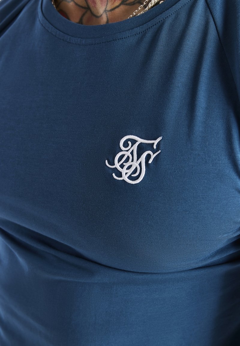 Gros plan sur une personne portant une chemise bleue avec un monogramme décoratif brodé en blanc sur la poitrine et un collier visible au-dessus du col.
