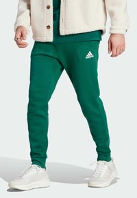 Green sweatpants with a fitted design featuring an embroidered white Adidas logo on the left hip, paired with a cream fleece jacket and white sneakers.