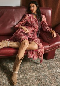 Vestito con motivo floreale in tonalità rosa con maniche lunghe, abbinato a stivali da cowboy color cuoio con cuciture decorative. Seduta su un divano bordeaux.