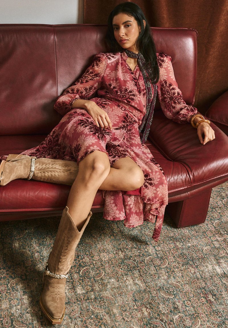 Vestito con motivo floreale in tonalità rosa con maniche lunghe, abbinato a stivali da cowboy color cuoio con cuciture decorative. Seduta su un divano bordeaux.