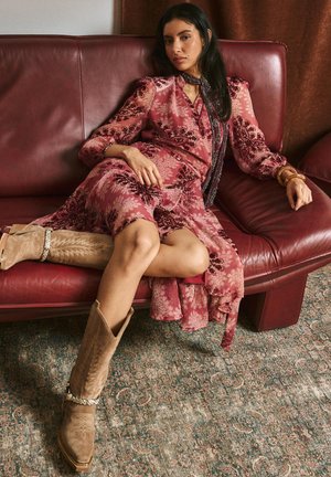 Kleid mit floralem Muster in Pinktönen und langen Ärmeln, kombiniert mit hellbraunen Cowboy-Stiefeln mit dekorativen Nähten. Sitzend auf einem bordeauxroten Sofa.