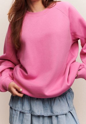 Personne portant un sweatshirt rose ample et une jupe en jean bleu clair à volants, une main dans une poche, haut du corps visible.