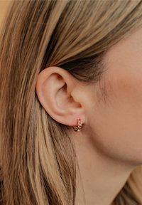 Petites boucles d'oreilles en or rose avec un design de maillons de chaîne, portées sur le lobe, mettant en valeur une texture lisse et un éclat subtil.