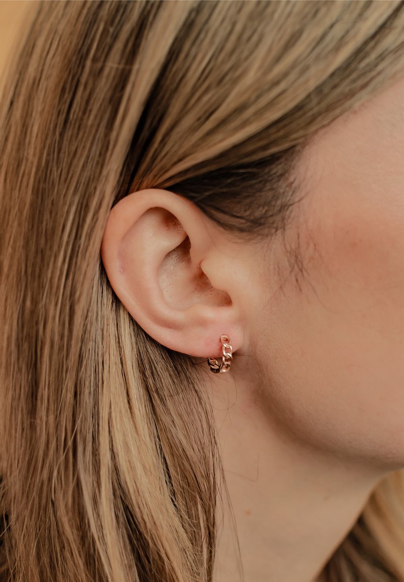 Petites boucles d'oreilles en or rose avec un design de maillons de chaîne, portées sur le lobe, mettant en valeur une texture lisse et un éclat subtil.