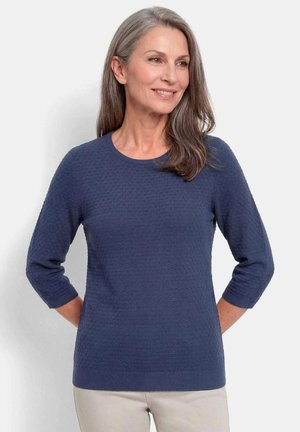 Frau mit grauen Haaren, die einen strukturierten dunkelblauen Pullover mit Dreiviertelärmeln und hellbeige Hosen trägt, lächelnd und seitlich schauend.