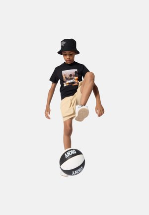 Schwarzes T-Shirt mit Grafikdruck, beige Shorts, weiße Sneakers und schwarzer Bucket-Hut. Junge Person balanciert einen schwarz-weißen Basketball.