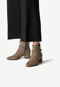 Bottines en daim taupe avec un talon bloc, fermeture éclair sur le côté et une sangle décorative avec des accents métalliques. Fini texturé.