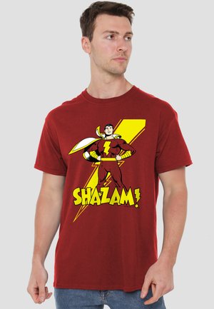 Young man wearing a red Shazam! superhero graphic t-shirt with yellow lightning bolt and text, standing against a plain gray background.