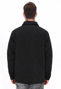 Zwarte jas met een gestructureerd patroon, puntige kraag, met knopen sluitbare manchetten en een ontspannen pasvorm. Zichtbare stiksels en zakken aan de achterkant.