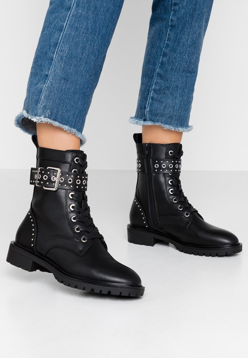 Bottes courtes en cuir noir avec laçage à l'avant, accents en métal argenté, sangle avec boucle et détails cloutés. Semelle épaisse et fermeture éclair latérale.