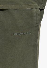 Gros plan d’un pantalon en tissu vert olive avec une poche latérale et un logo DROPSIZE en relief près de la poche.