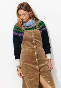Robe salopette en velours côtelé marron avec des boutons argentés et une fente sur le côté, superposée à un pull en laine coloré. Présente un poignet de chemise à carreaux.