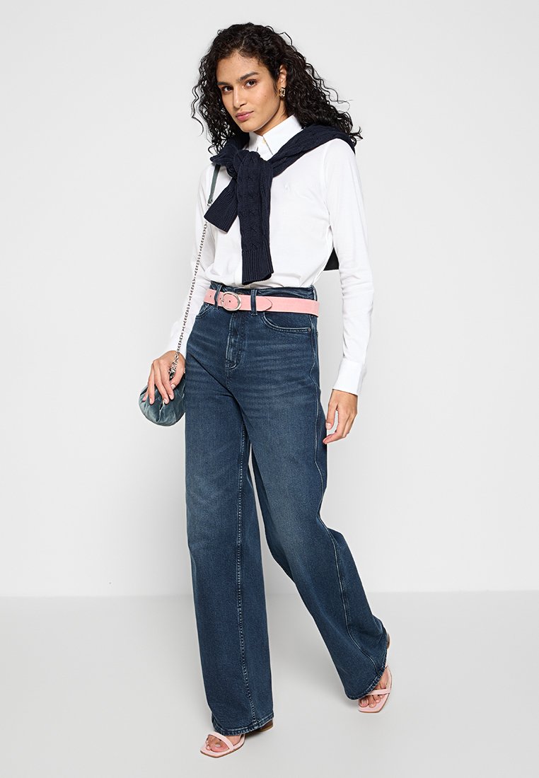 Jeans in denim scuro a gamba larga con una cintura rosa, camicia bianca con bottoni e un maglione blu navy draped sulle spalle. Porta una piccola borsa color turchese.