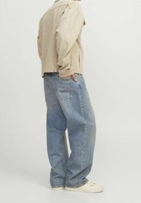 Beige överdimensionerad denimjacka med sidofickor, bärs över ljusblå baggy jeans. Modell som bär vita sneakers, sedd från sidan.