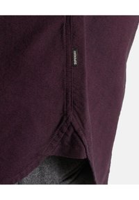 Tessuto viola scuro con una cucitura visibile e un piccolo etichetta nera con la scritta "SUPERDRY" vicino al bordo del tessuto.