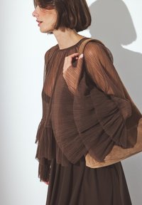 Blouse plissée marron avec des manches évasées et un col rond, associée à un sac à main en daim marron doux, sur fond minimaliste et épuré.