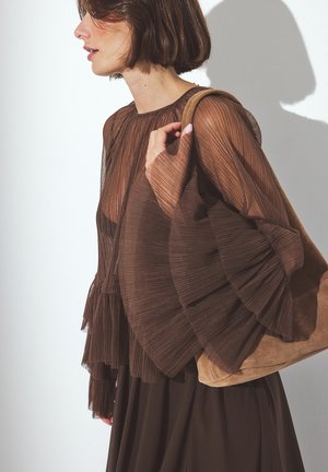 Bruine geplooide blouse met flared mouwen en een ronde halslijn, gecombineerd met een zachte bruine suède handtas, tegen een schone, minimalistische achtergrond.