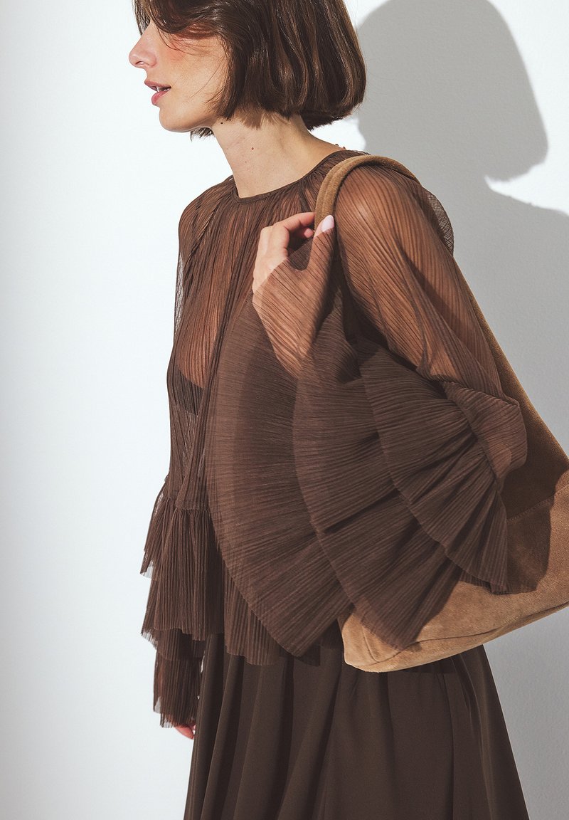 Blouse plissée marron avec des manches évasées et un col rond, associée à un sac à main en daim marron doux, sur fond minimaliste et épuré.