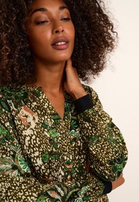 Blouse à manches longues à motif vert avec un décolleté en V, un devant à boutons et des accents de poignet noirs contrastants. Tissu texturé avec des motifs floraux.
