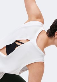 Personne portant un haut blanc sans manches avec un dos ouvert, révélant un soutien-gorge de sport noir en dessous, se penchant en avant avec un bras levé.