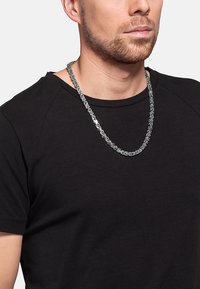 Sølvmetal kædelys med sammenflettede led, båret over en sort kortærmet skjorte. Glat overflade, blank finish, medium tykkelse.