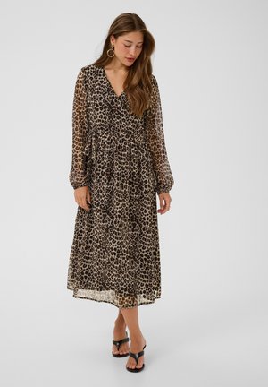 Kvinde iført en langærmet kjole med V-hals og leopardprint med en indsnørret talje, sorte åbne sko med hæl og creoler, stående på en ensfarvet baggrund.