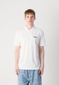 Hvit polo skjorte laget av bomull med en teksturert overflate, med krage og tre knapper. Svart "HUGO" logo på venstre bryst.