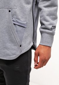 Grå hoodie med dragkedja, meshnätfickdetaljer och logotyp på ärmen. I kombination med mörka byxor med texturerad lagning.