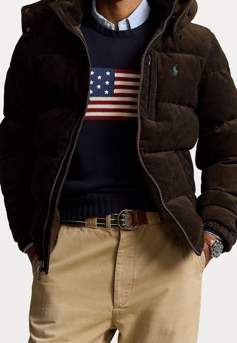 Veste matelassée en velours côtelé marron avec capuche, pull bleu marine avec un motif de drapeau américain rayé rouge et blanc, chinos beige et ceinture en cuir.