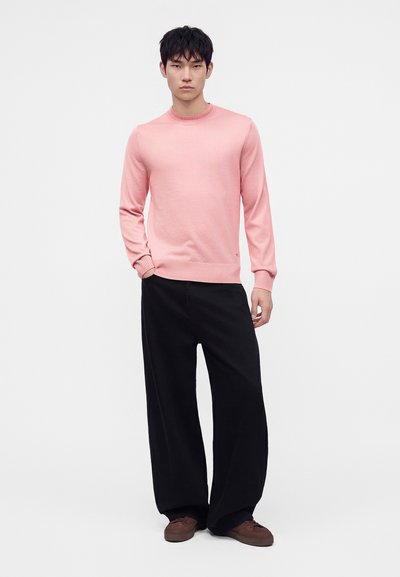 Jeune homme portant un pull rose, un pantalon large noir et des chaussures marron, debout avec une main dans la poche devant un fond uni.