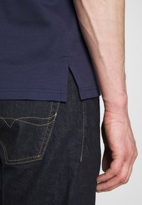 Námořnická modrá bavlněná tričko s bočním rozparkem, spárováno s tmavými džínami s zlatým prošíváním na kapsách a švech.