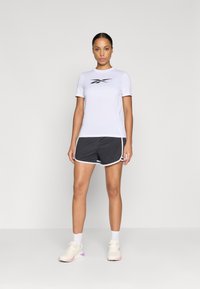 Tricou athletic Alb cu logo negru, asortat cu pantaloni scurți atletici gri închis cu dungi albe și adidași deschiși la culoare.