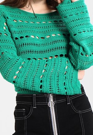 Femme portant un haut vert en crochet à manches longues et un pantalon noir taille haute avec une fermeture éclair argentée à l'avant et des coutures blanches.