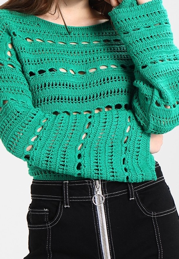 Femme portant un haut vert en crochet à manches longues et un pantalon noir taille haute avec une fermeture éclair argentée à l'avant et des coutures blanches.