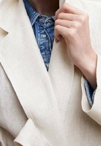 Lys beige linned blazer med spids flip, lagd oven på en blå denimskjorte, der viser et struktureret stof og en enkelt knapdetalje.