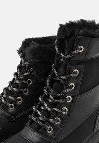 Black boots featuring a faux fur cuff, leather and textile upper, silver eyelets, and striped laces with a textured finish.