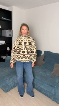 Kremowy polarowy pulower z brązowymi geometrycznymi wzorami, wysokim kołnierzem i ćwiartkowym suwakiem; w połączeniu z niebieskimi rozszerzanymi dżinsami i trampkami.